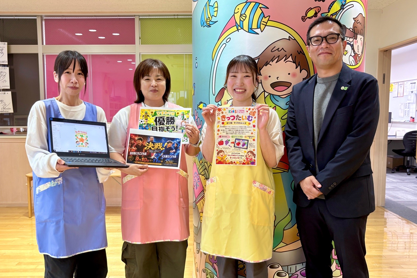 ヒナリーで作った掲示物とパソコンを手に並ぶ、東月寒にれこども園の先生方と園長